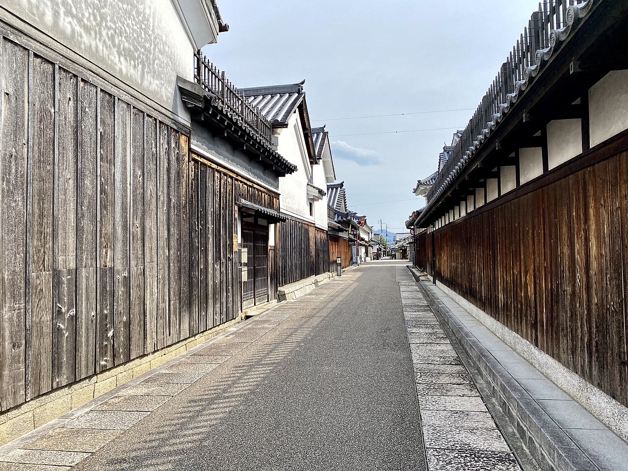 A quiet lane in the Jinaimachi merchant quarter of Tondabayashi, lined with weathered wooden fences and white-walled storehouses under tiled roofs