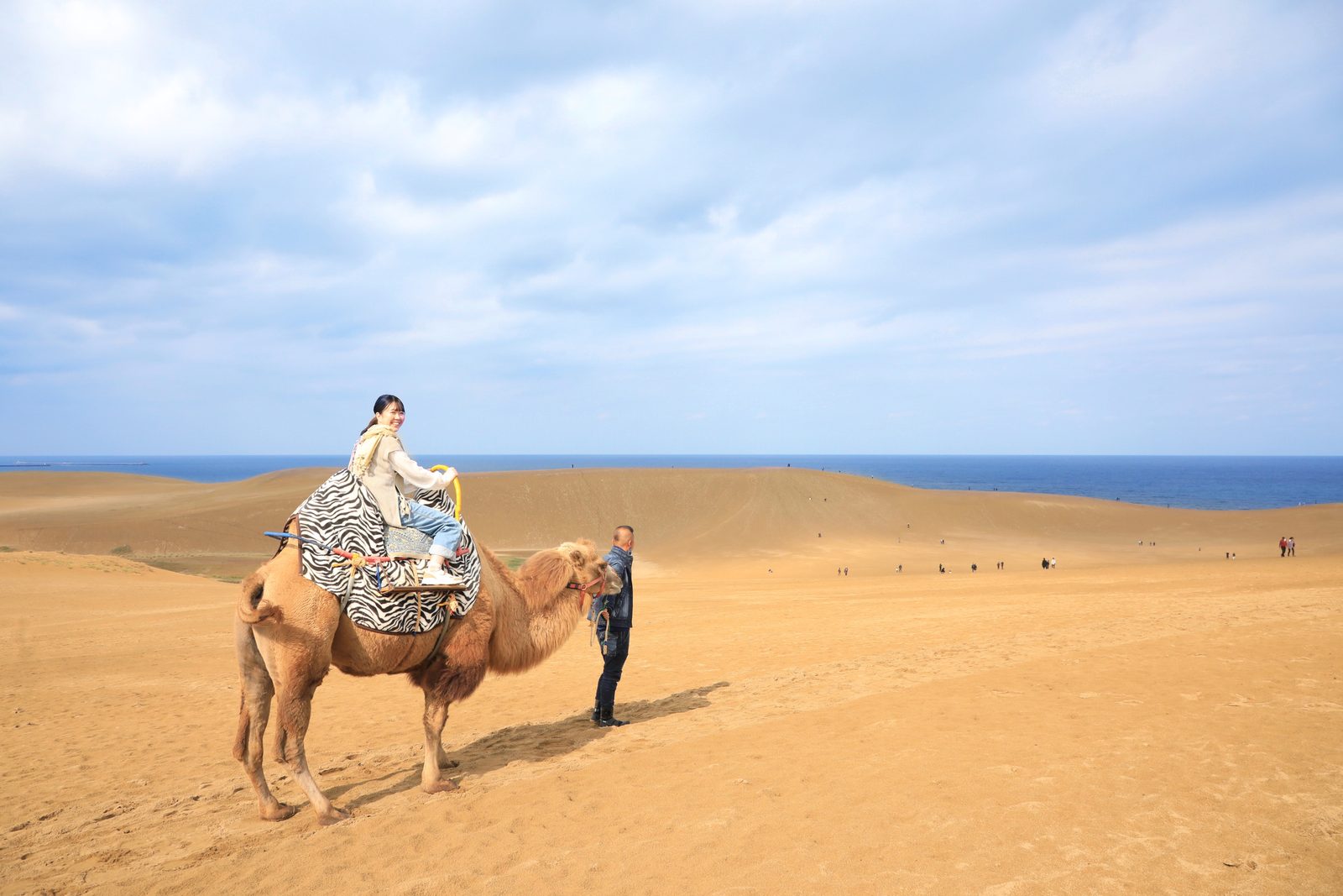 A visitor riding a camel on the dunes led by a handler, blue sea on the horizon