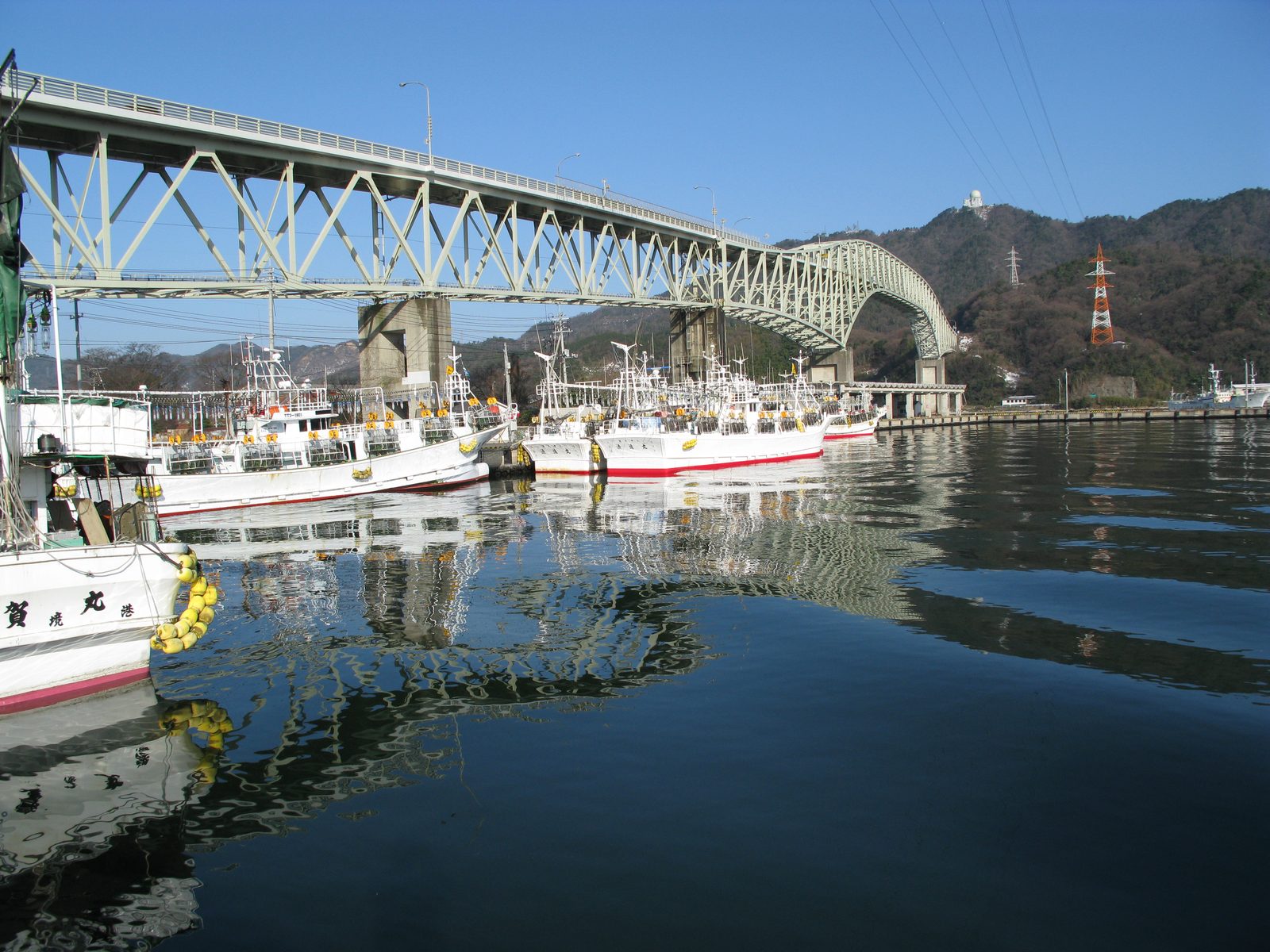 White fishing boats moored in a harbour under a steel arch bridge, with the boats and bridge reflected in dark water and hills behind