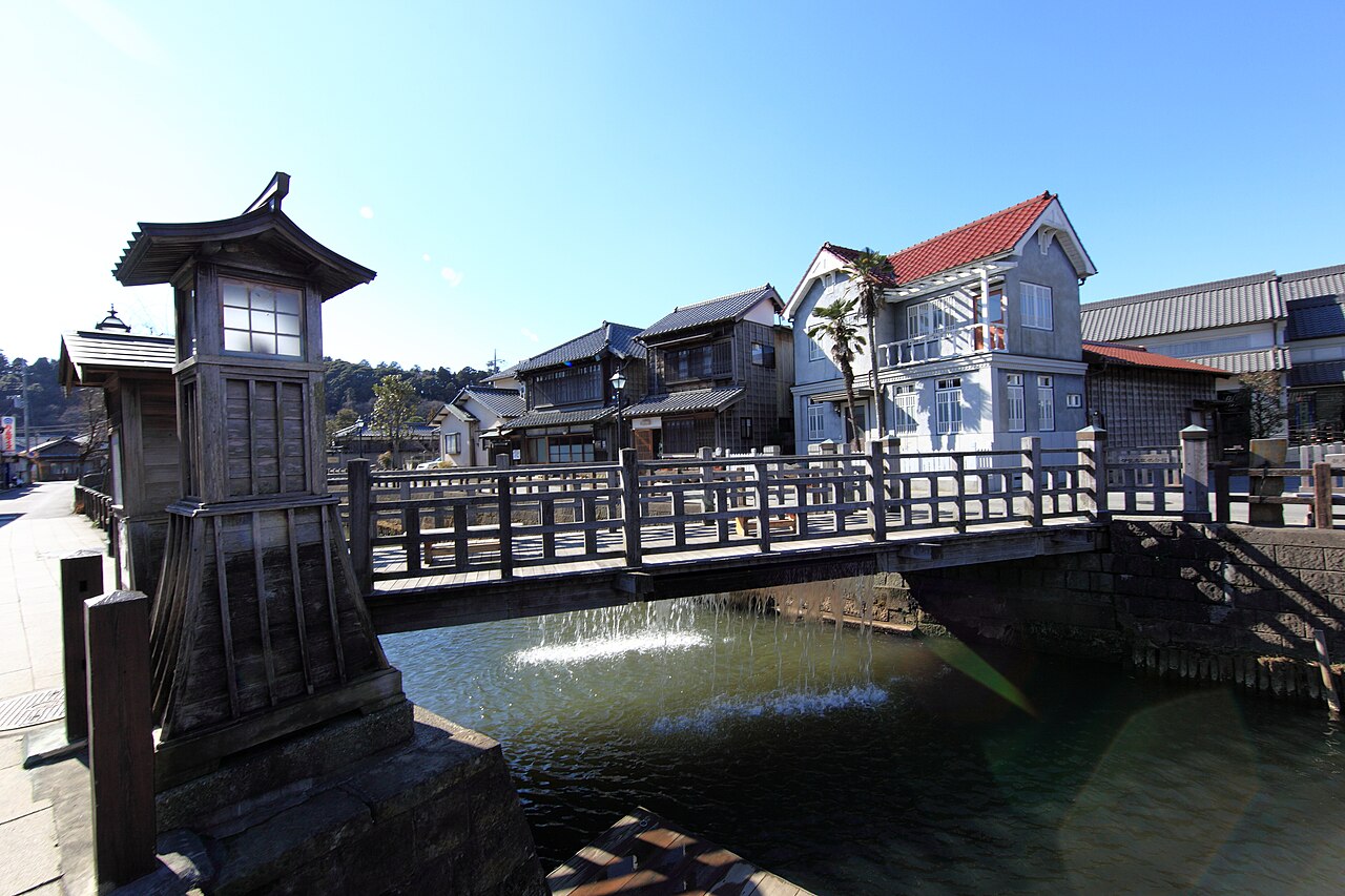 The wooden Toyohashi bridge over the Ono River in Sawara, water cascading from its underside weir past a lantern-topped lookout post and old townhouses along the bank.