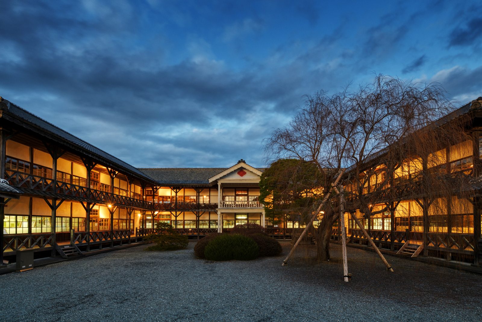 Two-story wooden former school building with a white balcony and a gravel courtyard at dusk.