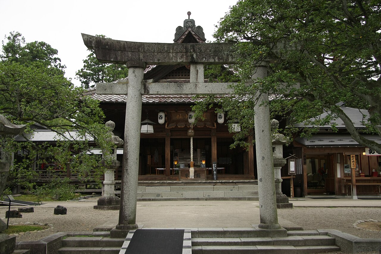 Shonai Shrine inside Tsuruoka Park, framed by a grey stone torii, with the wooden worship hall and its paper lanterns behind it and old trees leaning over the gravel forecourt.