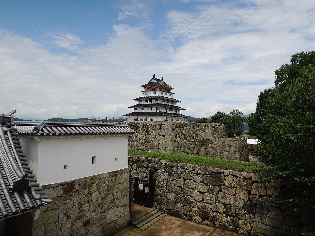 A reconstructed castle tower at Tsuyama Castle rising behind a whitewashed gate wall, seen over the site's high stone ramparts