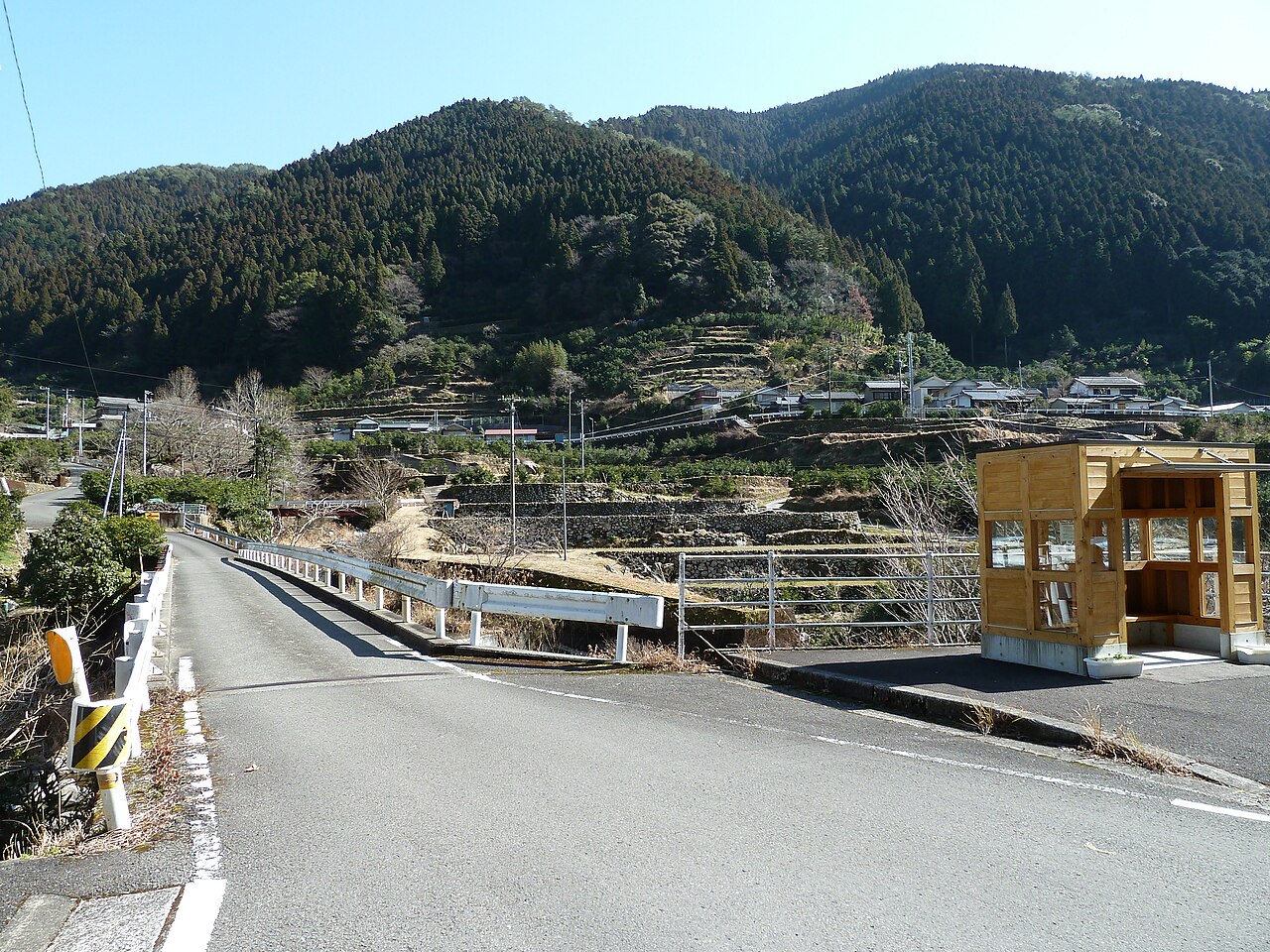The entrance road into Umaji Village, an empty lane passing terraced fields with cedar-covered mountains rising behind the houses.