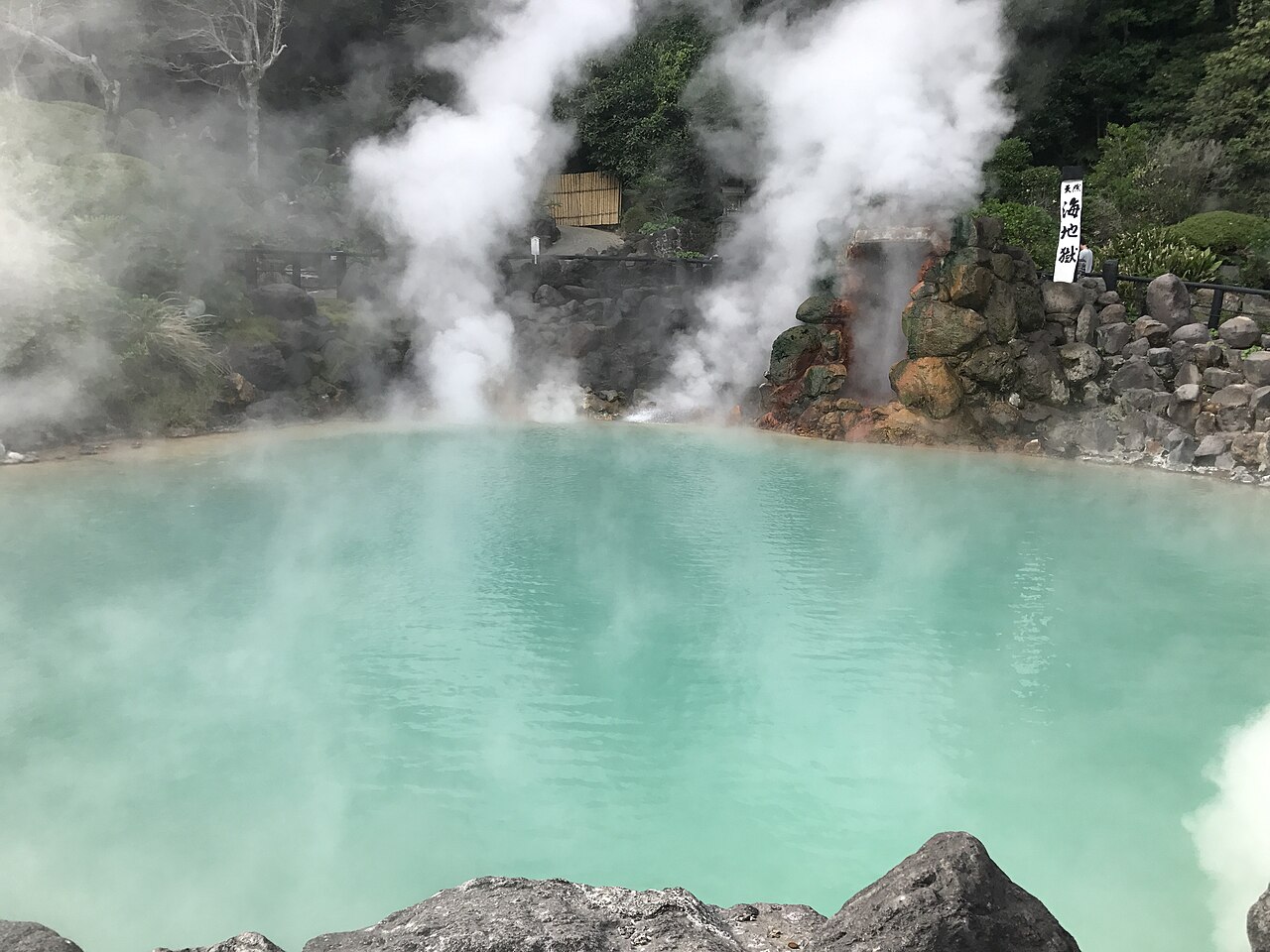 Steam rising from the milky turquoise pool of Umi Jigoku in Kannawa, Beppu, with a stone-walled bank and a vertical signboard behind it.