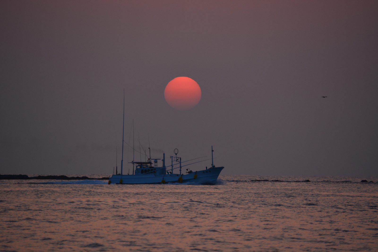 A fishing boat crosses a calm sea beneath a round red sun hanging low in a hazy sky
