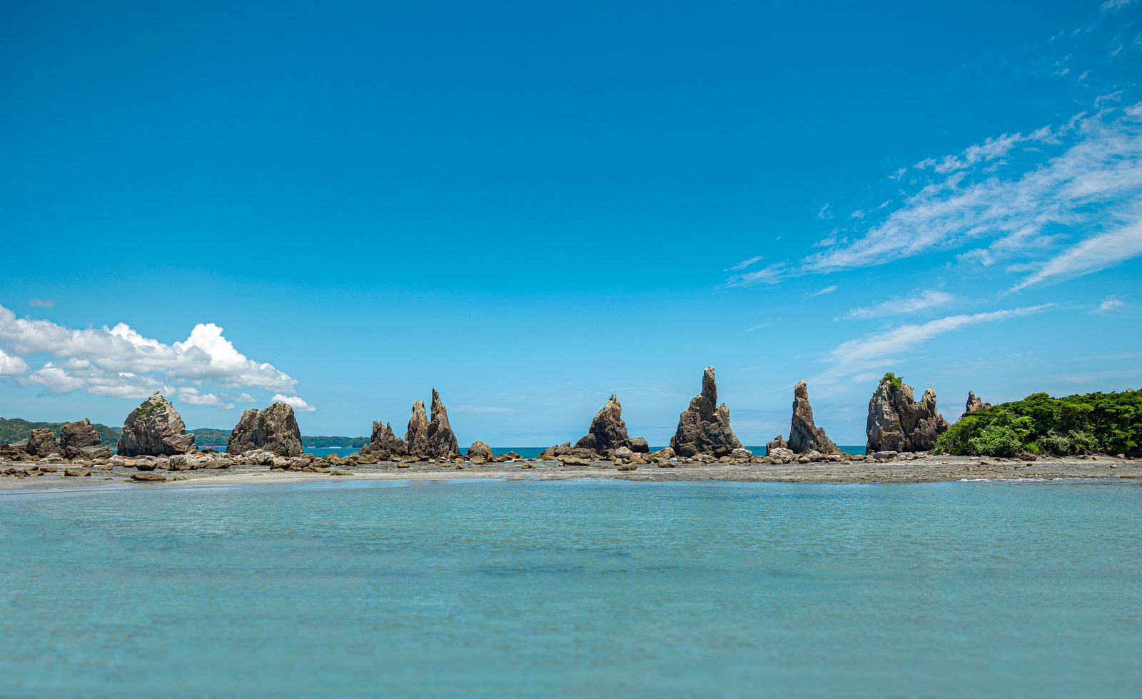 A row of jagged rock spires stands along a low shoreline across shallow turquoise water, under a deep blue sky with thin white clouds