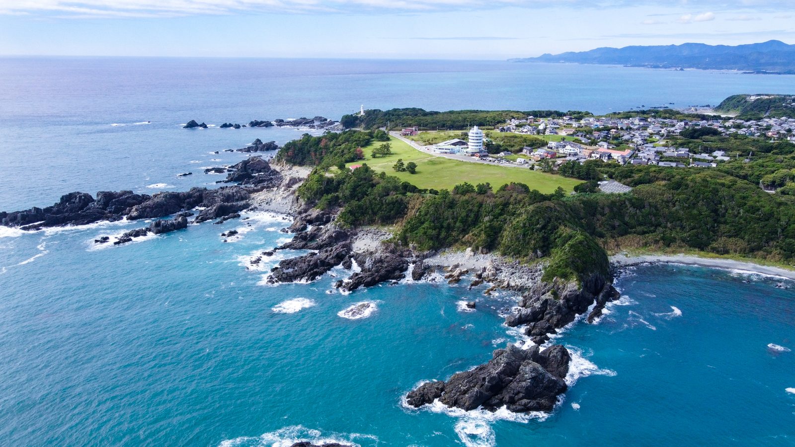 Seen from above, a green headland of grass and low trees juts into a blue sea with black rocks and white surf, a white round building and a town beyond