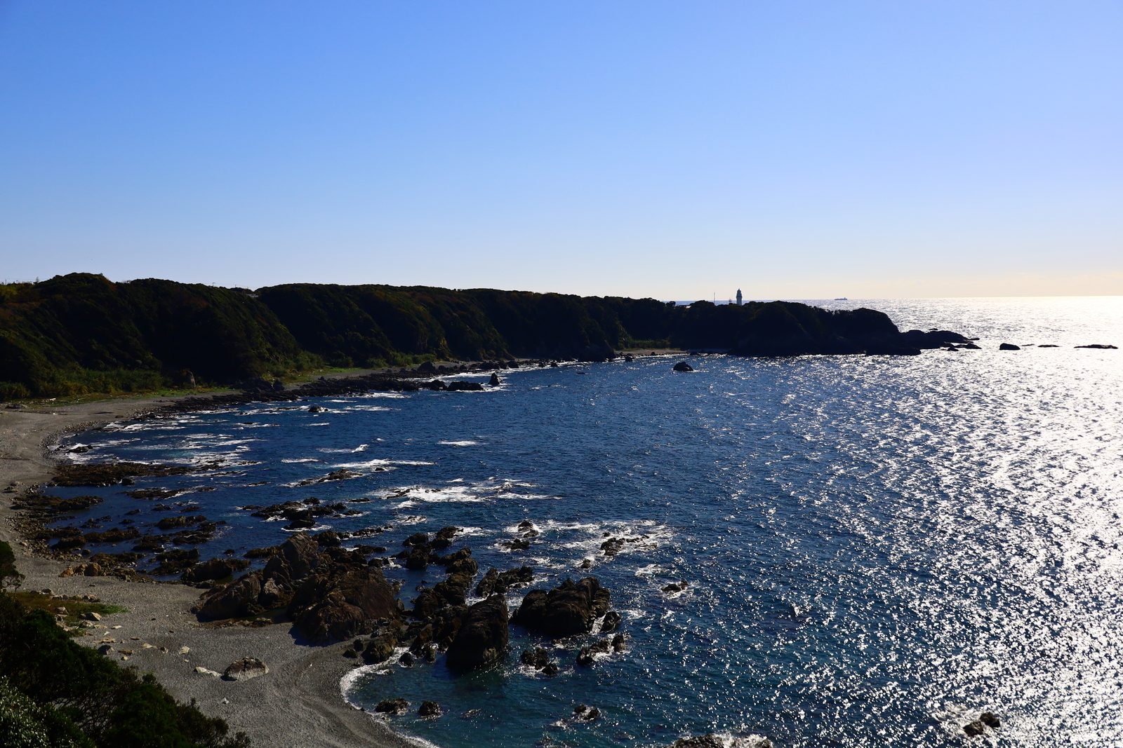 A rocky cove with a gravel beach curves toward a dark wooded headland, a small white lighthouse on the far point, and sunlight glittering across the dark blue sea