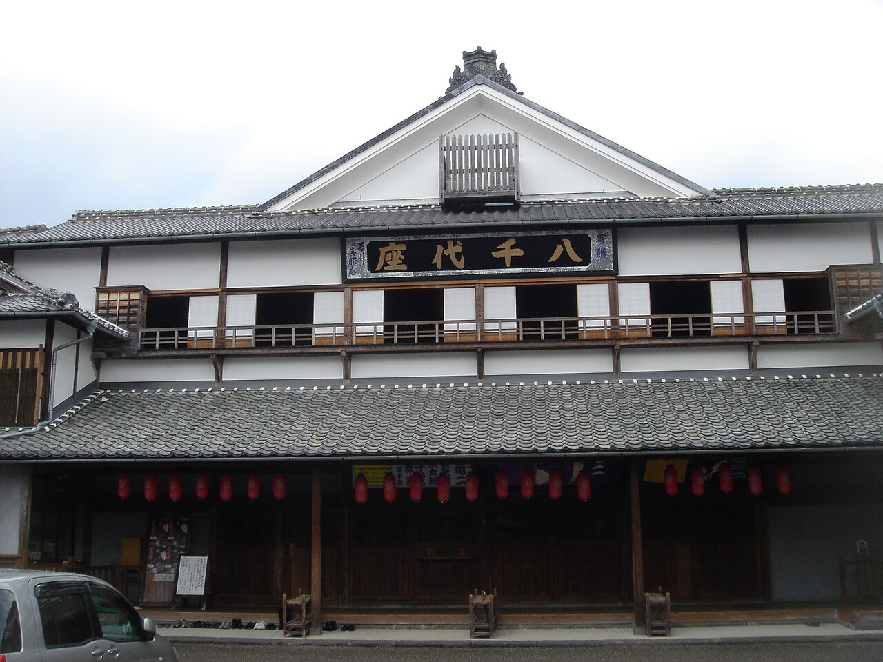 The wooden two-storey street front of the Yachiyo-za theatre in Yamaga, with a row of red paper lanterns hanging under the tiled eaves below a gilded signboard.