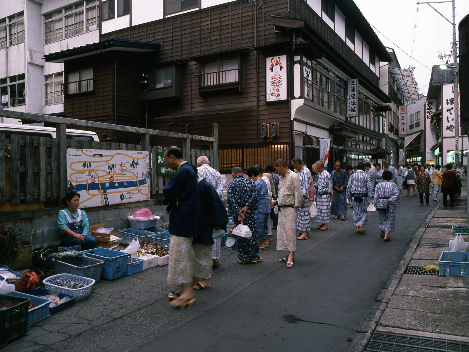 Guests in yukata walking and browsing along a narrow hot-spring street where a woman sits beside baskets of vegetables, with wooden inns and shop signs behind