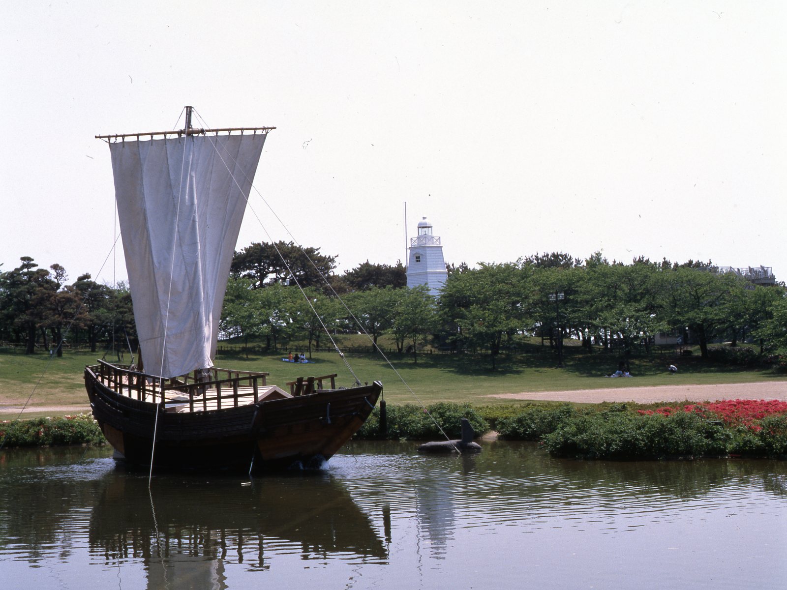 Wooden sailing ship with white sails floating on a pond in Hiyoriyama Park, with the white six-sided lighthouse in the distance