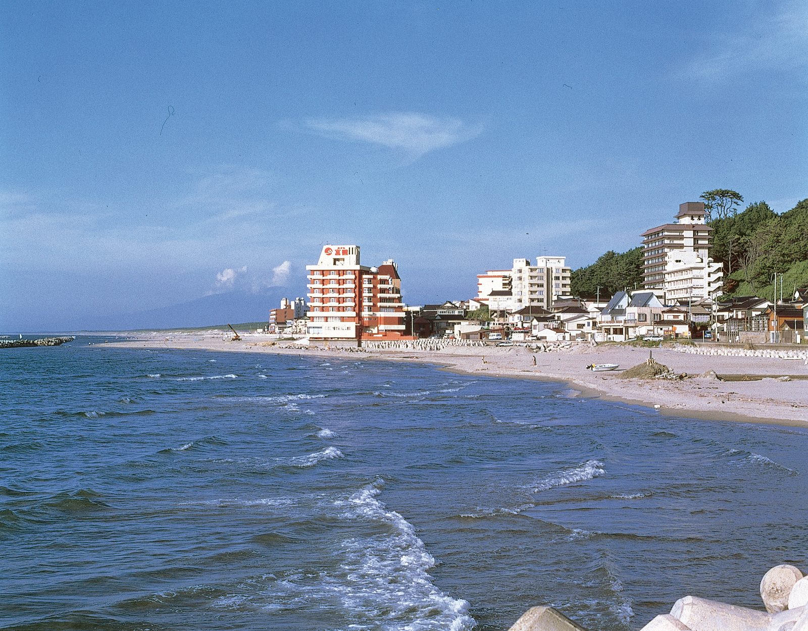 Gentle waves rolling onto the sandy beach at Yunohama Onsen, with a row of inns and hotels along the shore below a wooded hillside and a blue sky.