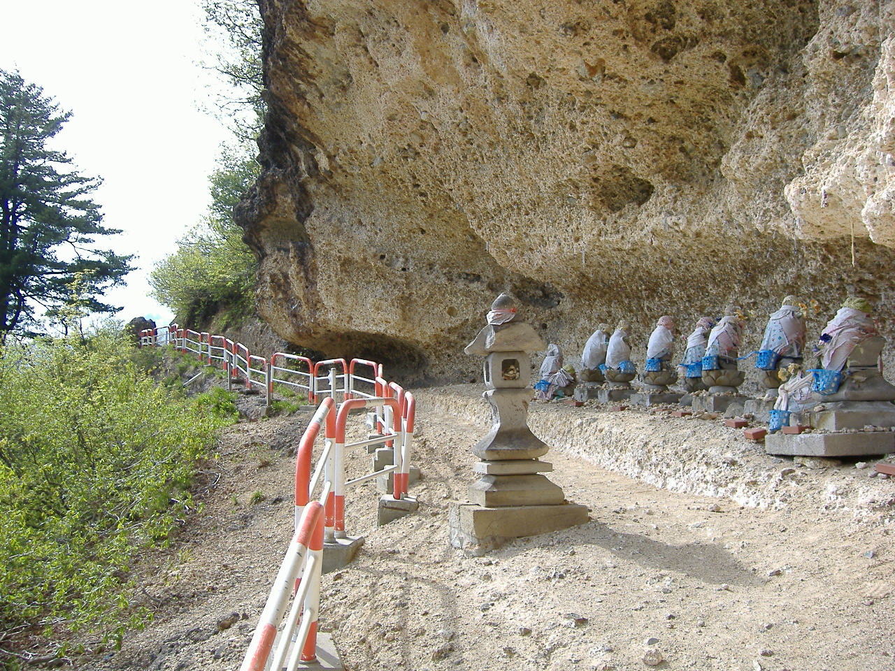A row of small stone Jizo figures in blue and red cloth beneath a huge overhanging rock face, with stone lanterns and a rail along the dirt path