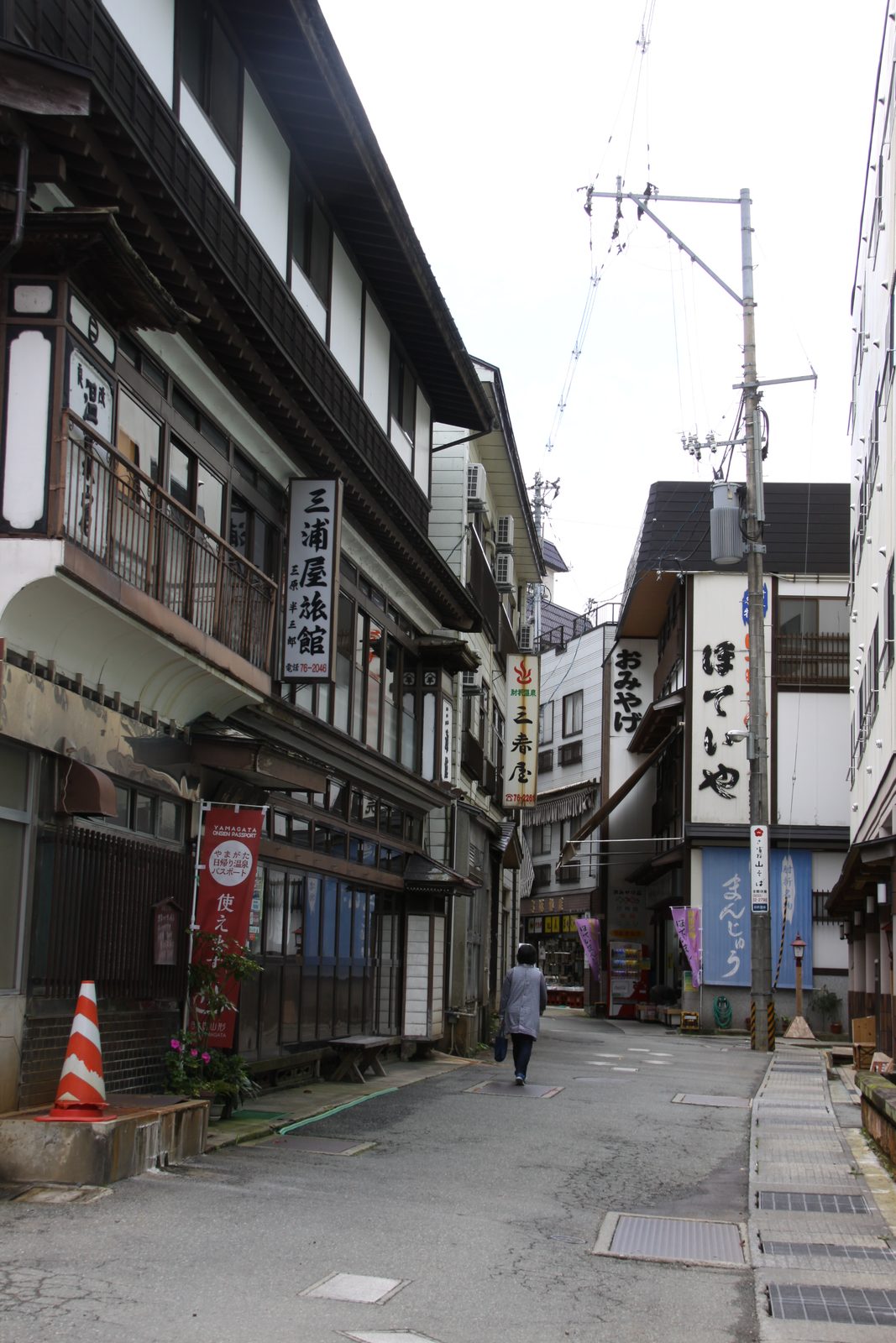 A quiet empty street in an old hot-spring town, with multi-storey wooden inns on both sides, utility wires overhead and a single walker far down the road