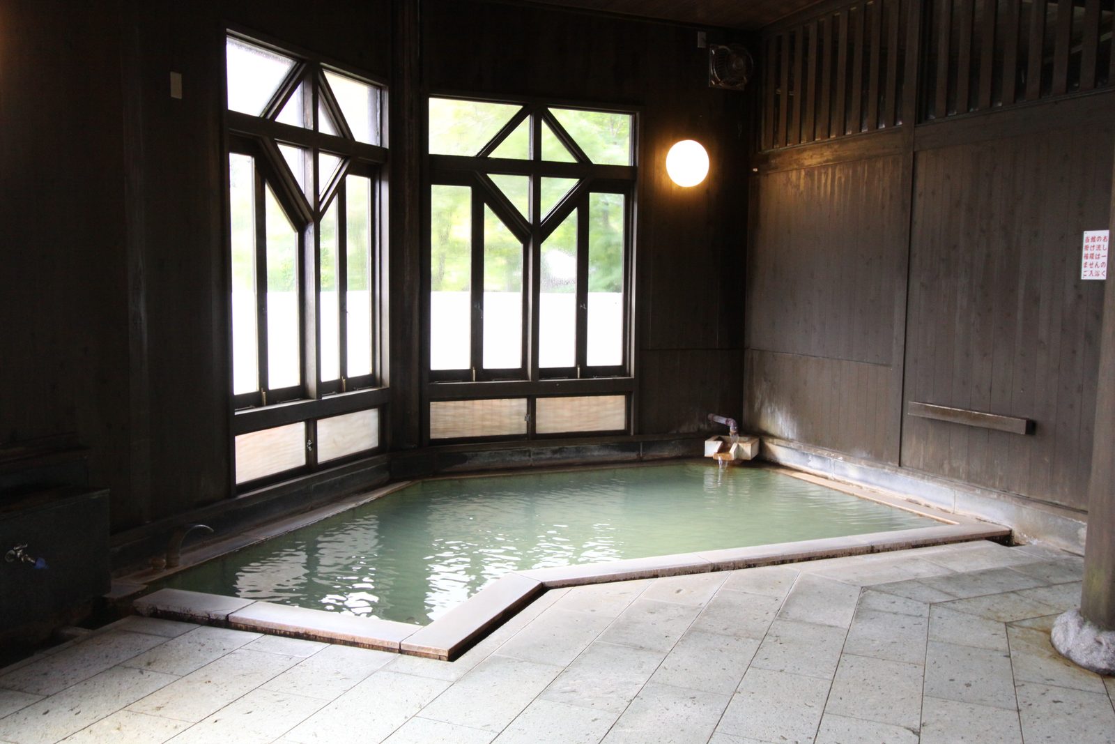 Old wooden bathhouse interior with a stone-edged pool of pale milky-green water and a tall window with geometric lattice in the corner