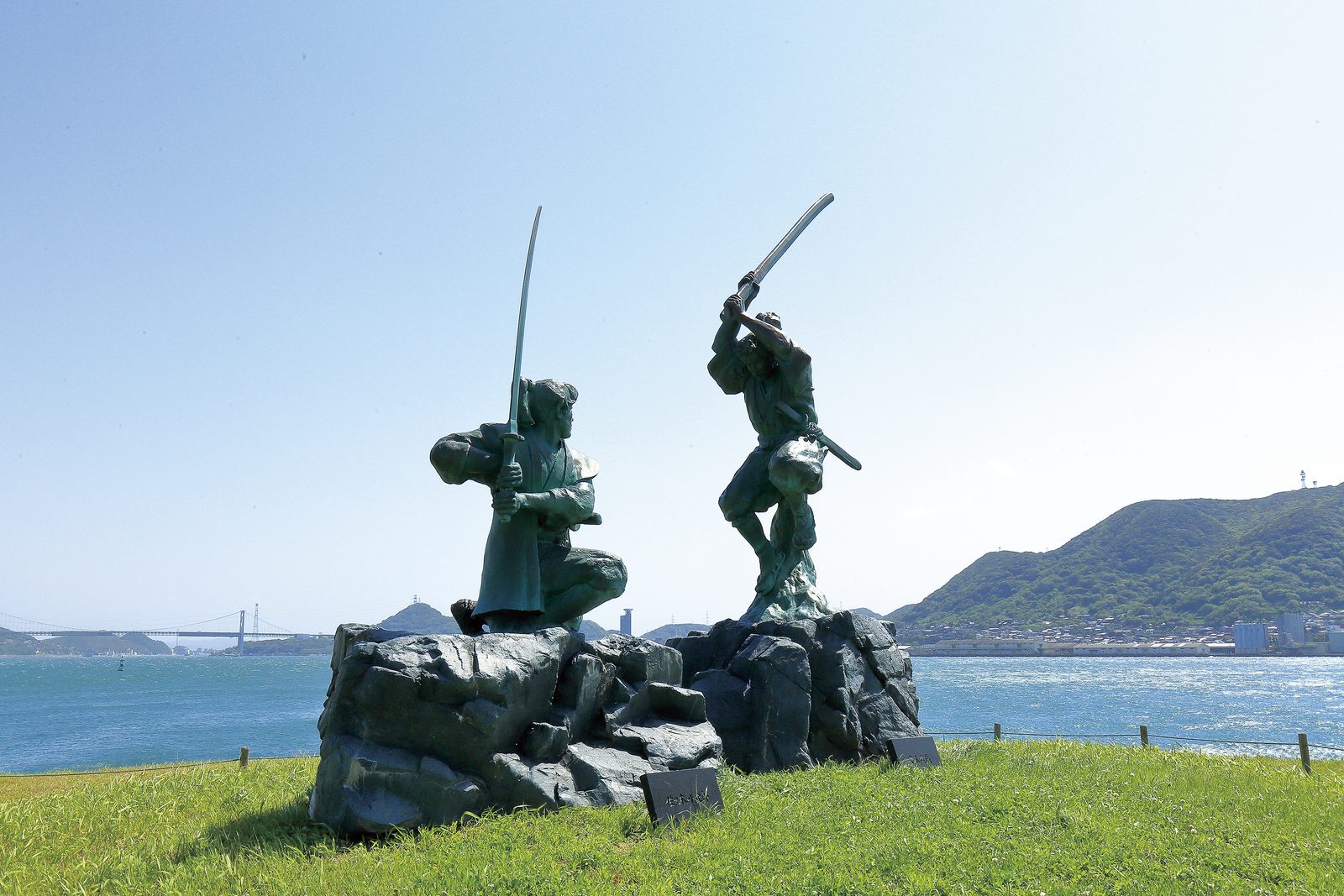 Bronze statues of two duelling swordsmen on the grass of Ganryujima, with the strait behind