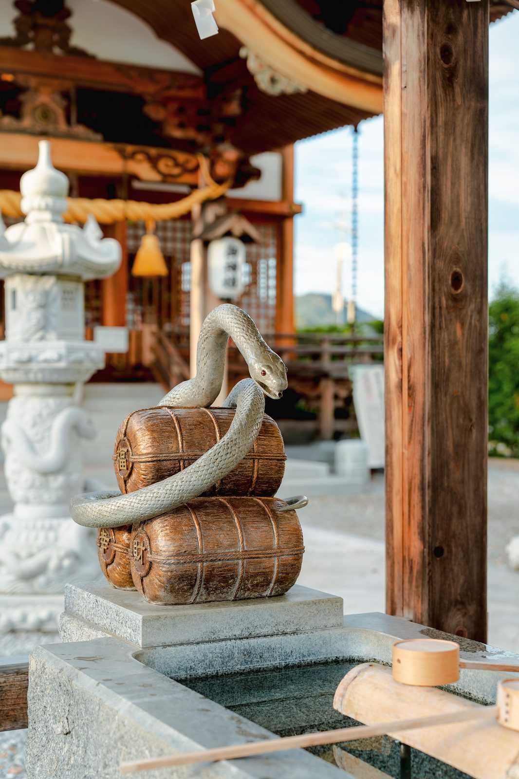 A white snake statue coiled over rice bales beside a shrine water basin in Iwakuni