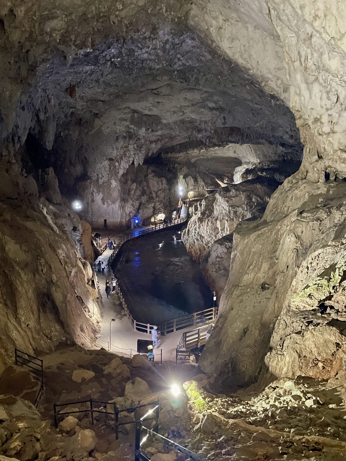 A walkway with handrails following a dark underground pool inside Akiyoshido Cave, with small figures of visitors far ahead