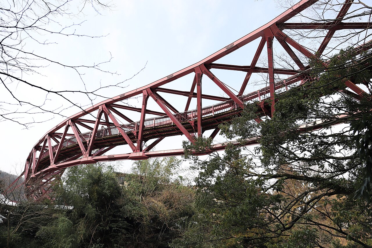 A steel truss footbridge with an intricate openwork frame spans the wooded Kakusenkei gorge at Yamanaka Onsen, seen from beside the stream below.