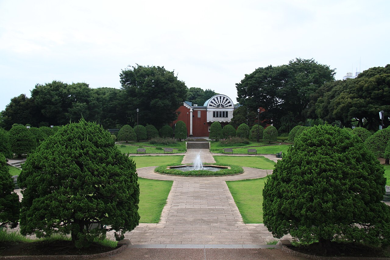 A fountain and clipped hedges lead across a lawn plaza toward a two-storey brick building with a fan-shaped window, in the Yamate district of Yokohama.