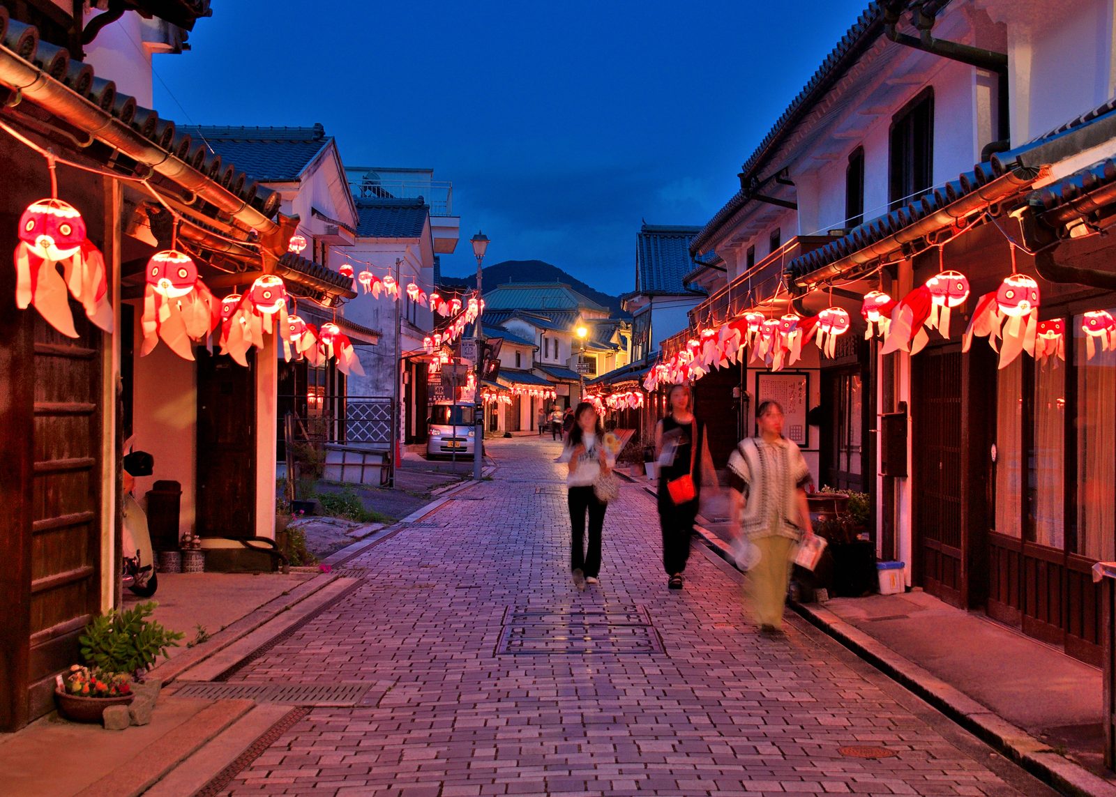 Cobbled street lined with white-walled merchant houses at dusk, hung with glowing red goldfish lanterns, with three people walking toward the camera and a hill beyond