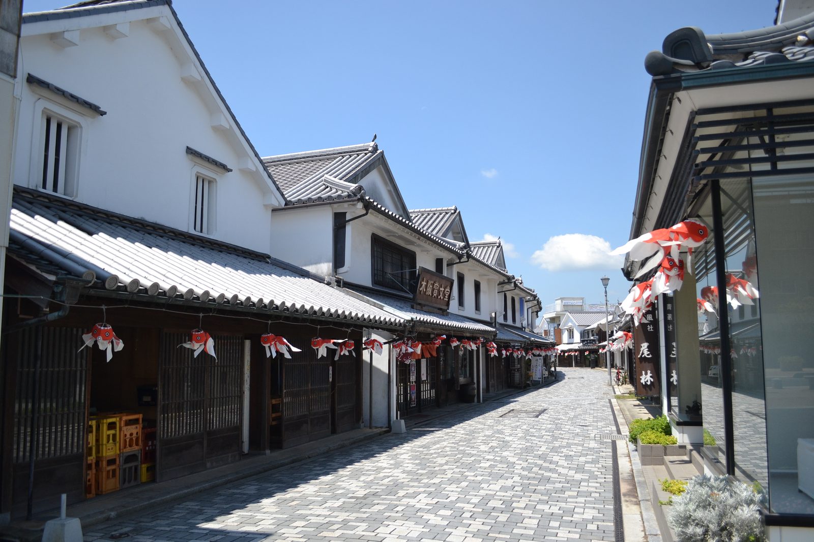 Daytime view down the stone-paved white-wall street in Yanai, with whitewashed storehouses with tiled roofs on the left and small goldfish lanterns hanging under the eaves