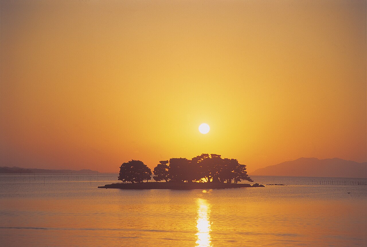 Yomegashima, a small pine-covered island in Lake Shinji, silhouetted against an orange evening sky with the sun low over the water.